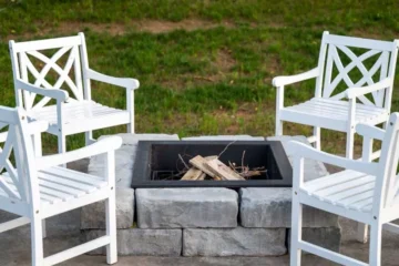 A stone fire pit with a square insert surrounded by 4 white patio chairs - How to build a fire pit